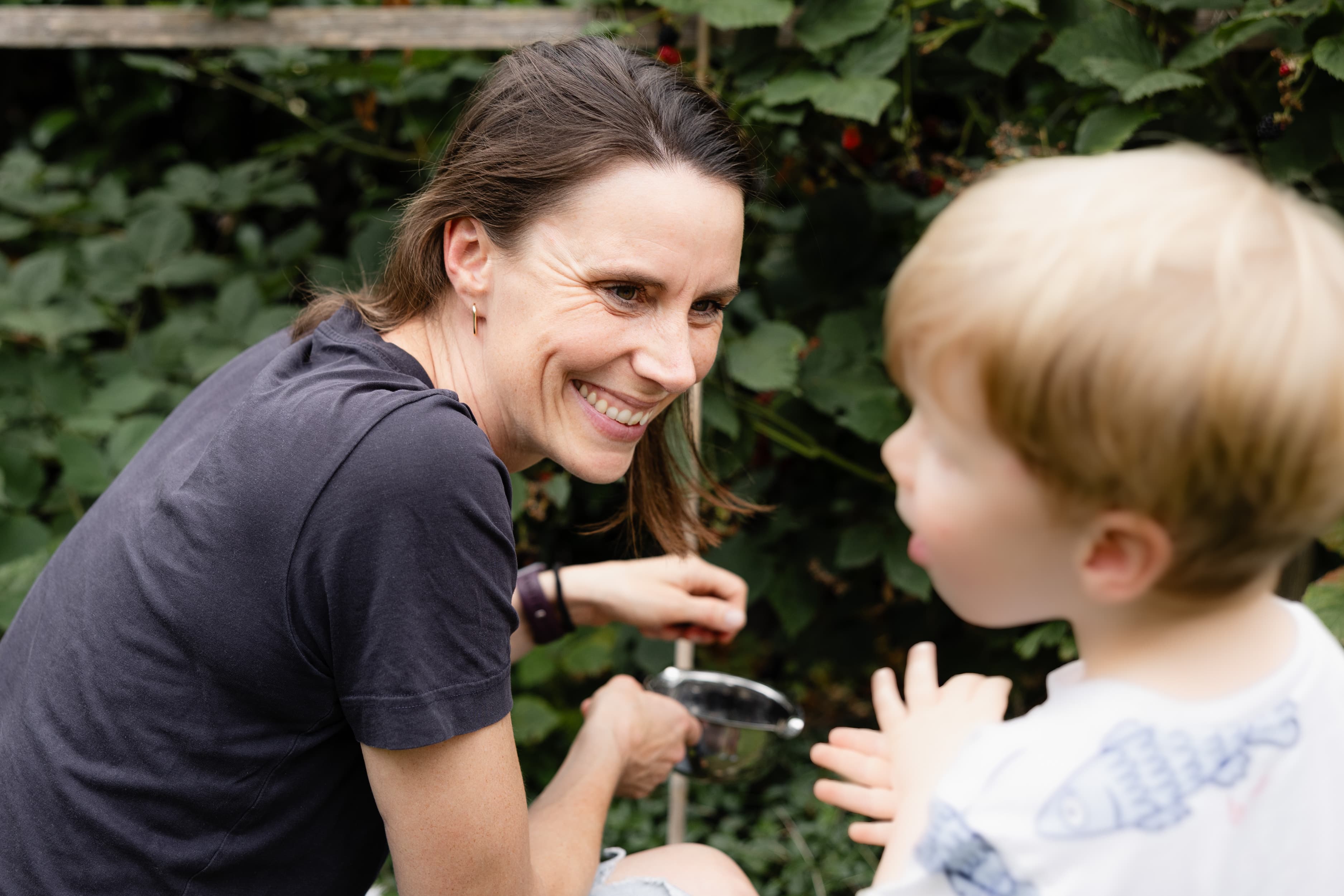 Flexible Arbeitszeitmodelle Eine Mitarbeiterin pflückt mit ihrem kleine Sohn Brombeeren im Garten. Sie lächelt ihren Sohn an, der unscharf im Vordergrund zu erkennen ist.